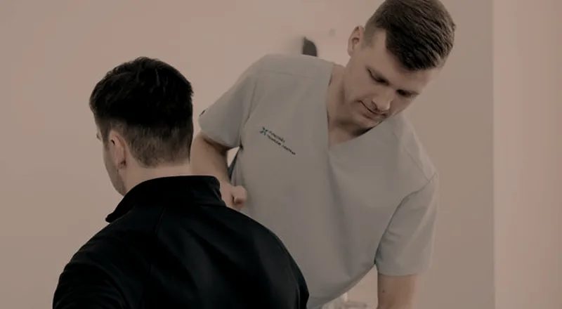 Profissional de saúde examinando o ombro de uma pessoa em pé, em sala de atendimento.