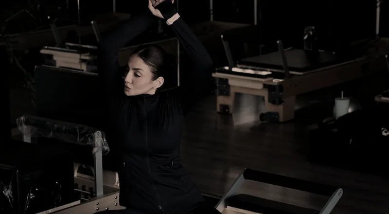 Mulher sentada em um reformer, braços estendidos acima da cabeça, em studio com vários aparelhos.