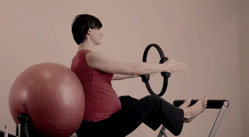 Gestante apoiada em bola suíça, segurando um arco de pilates com os braços à frente.